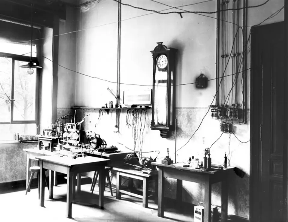 Siyah-beyaz fotoğrafta masalarında deney düzenekleri bulunan ve duvarında sarkaçlı saat asılı eski bir laboratuvar